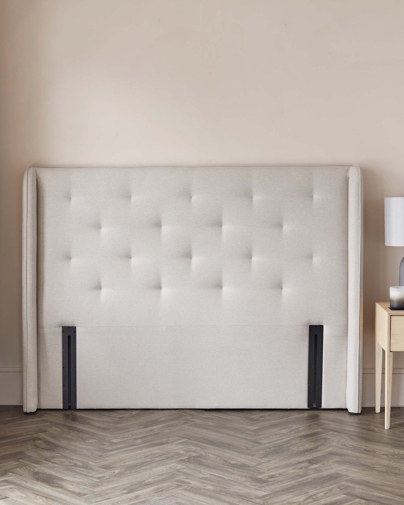 Elegant upholstered king-sized bed headboard in a light neutral colour with diamond tufting and clean, straight edges. Contemporary wooden bedside table with a drawer stands next to it, featuring a simple lamp atop.