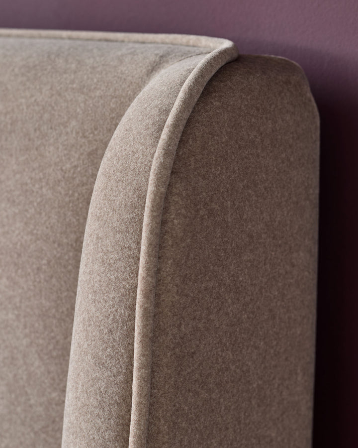 Close-up of a light brown upholstered headboard with a curved top and piped edge, against a dark red wall.