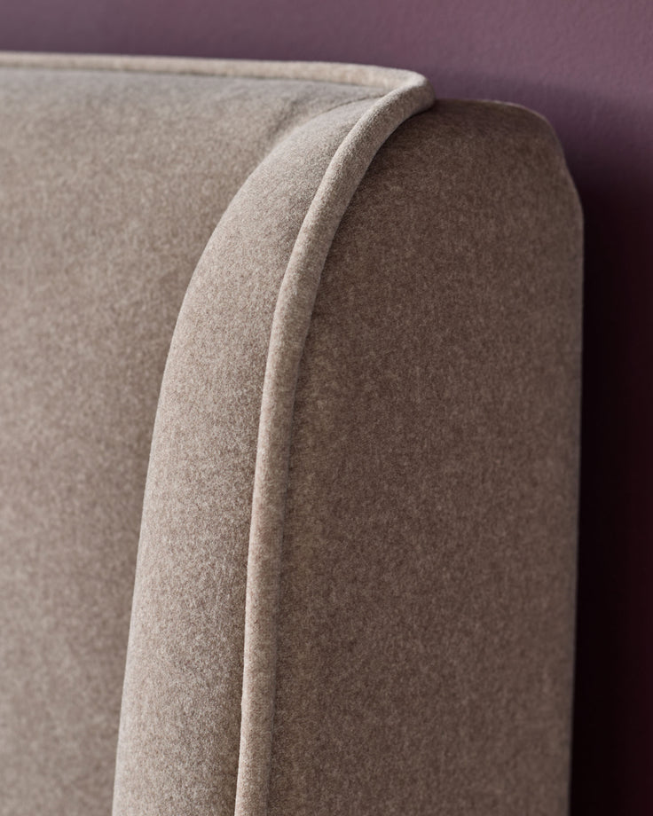 Close-up of a light brown upholstered headboard with a curved top and piped edge, against a dark red wall.