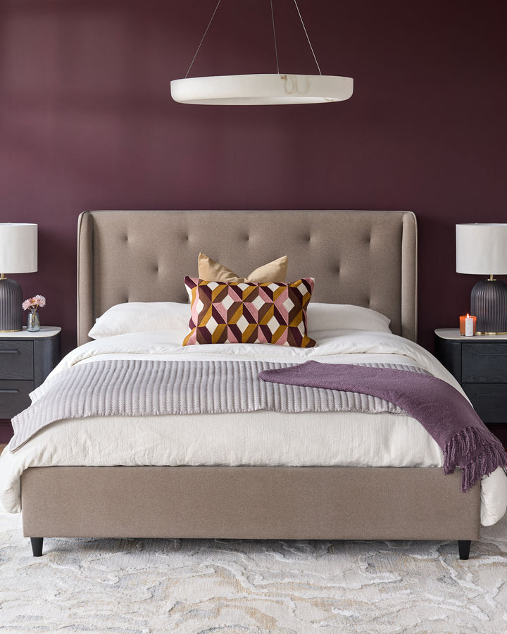 A modern bedroom with a taupe upholstered bed, bedside tables with lamps, and geometric pillows.