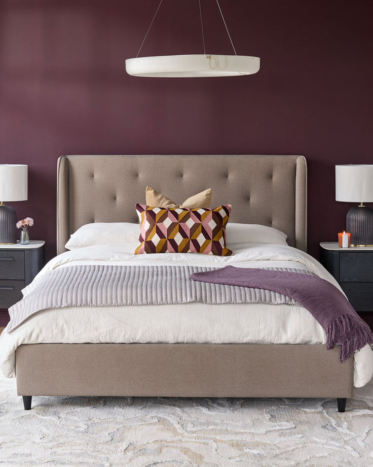 A modern bedroom with a taupe upholstered bed, bedside tables with lamps, and geometric pillows.