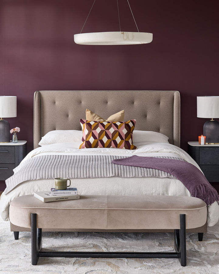 Bedroom with neutral-toned bed, bench, bedside tables and lamps. Accent colours include plum and gold.