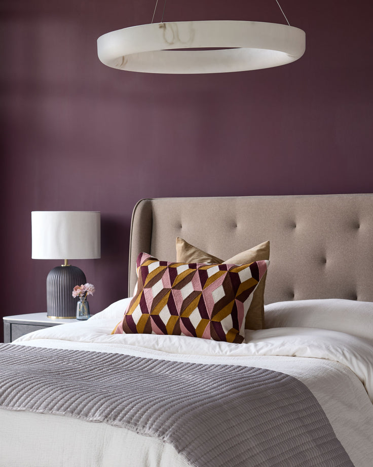 Bedroom with bed, upholstered headboard, throw, cushions, lamp and circular ceiling light. Rich plum wall colour.