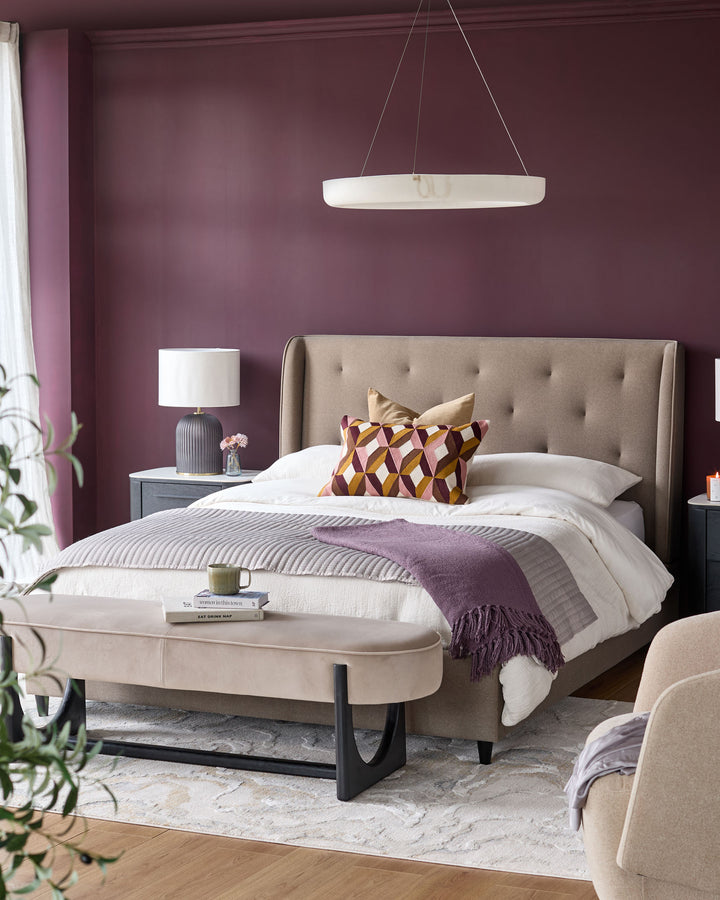Bedroom with a beige upholstered bed, bench and accent chair, on a cream-coloured patterned rug. Walls are a dark plum colour.