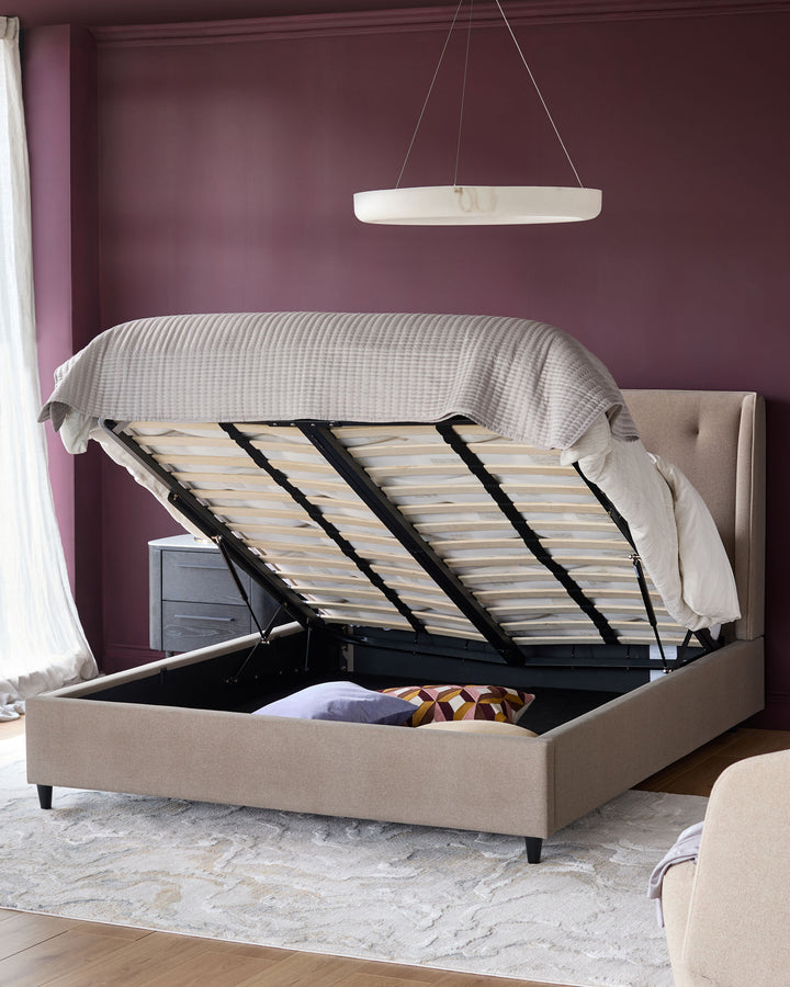 Beige ottoman bed with lift-up frame and a modern hanging light fixture, set against a plum-coloured wall.