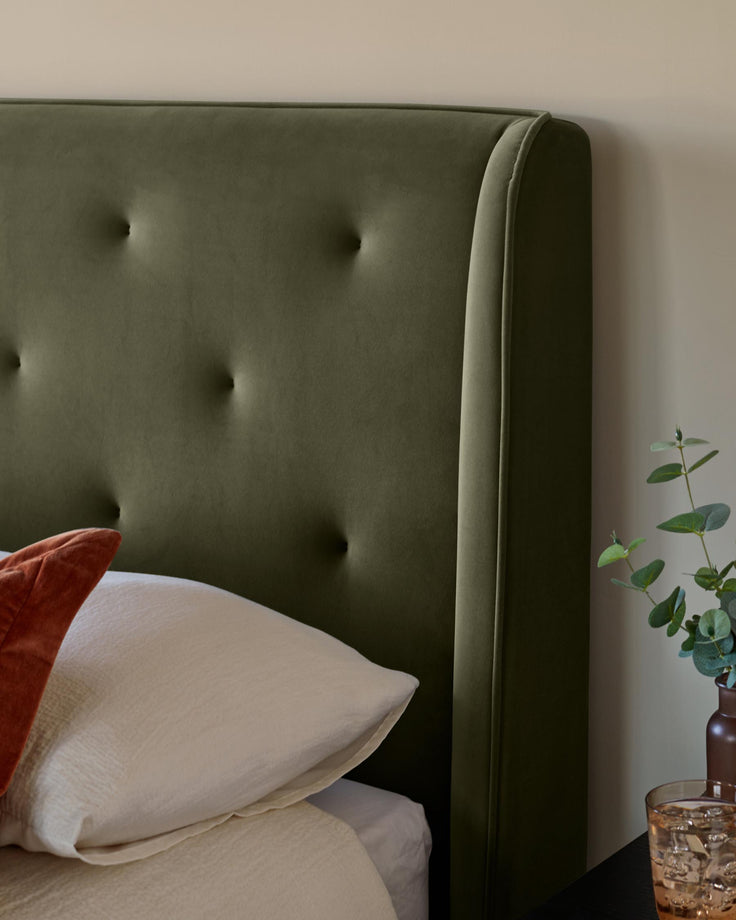 Olive green velvet headboard with button detailing. Pillows sit on a light-coloured duvet. Eucalyptus in a vase is to the side.