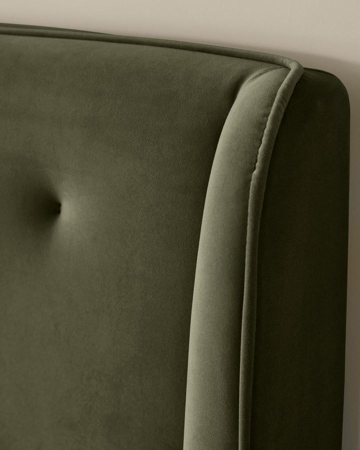 Close-up of an olive green upholstered headboard with a button-tufted design and piped trim. Soft, textured fabric.