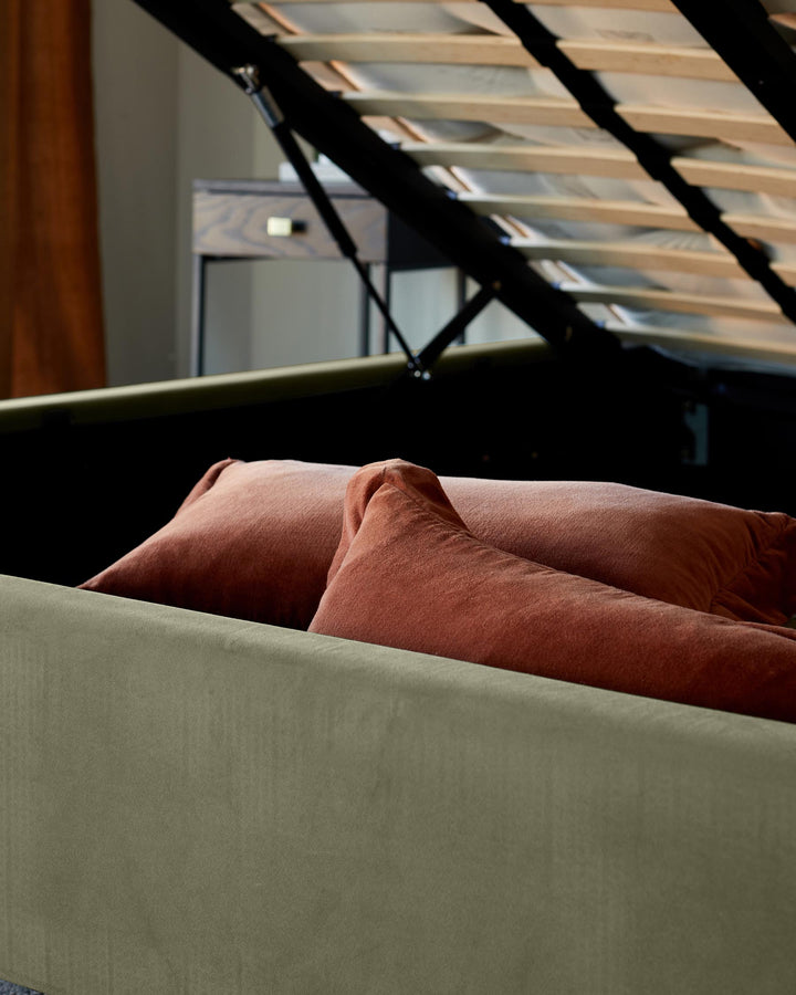 A lift-up ottoman bed with reddish brown pillows, revealing storage space and a bedside table in the background.