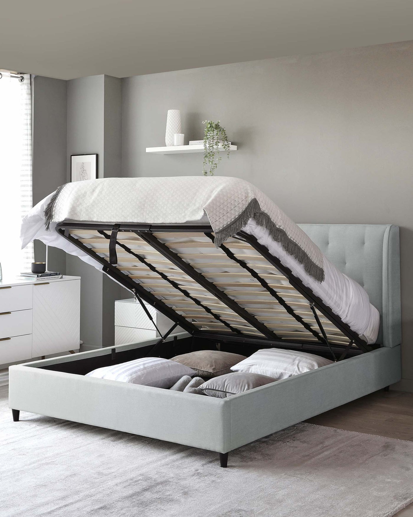 A modern light grey upholstered storage bed with its mattress lifted to reveal a spacious storage compartment underneath. The bed features a tufted headboard and is set on dark wooden legs. A matching light grey nightstand with white drawer fronts is visible to the left of the bed.