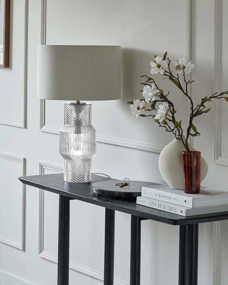Elegant black console table with a textured finish, featuring slender, tapered legs and a smooth top surface, accessorized with a modern, clear ribbed glass lamp with a white shade, a white vase with cherry blossoms, and a stack of decorative books.
