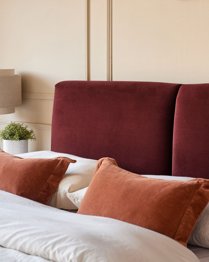 Leona super king ottoman bed in burgundy velvet with rust-colored pillows and cream bedding, shown at foot-of-bed angle.