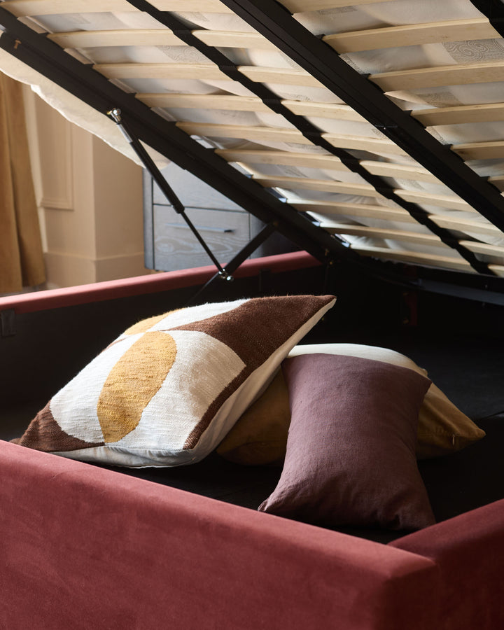 Storage bed with red velvet-like upholstery, revealing a hidden storage compartment with throw pillows.