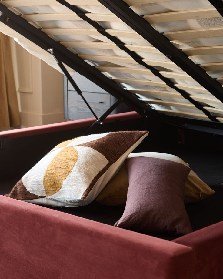 Storage bed with red velvet-like upholstery, revealing a hidden storage compartment with throw pillows.