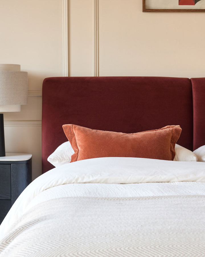 Cosy bedroom scene with a plush red headboard, complementary orange pillow, and white bedding. A bedside table is visible.