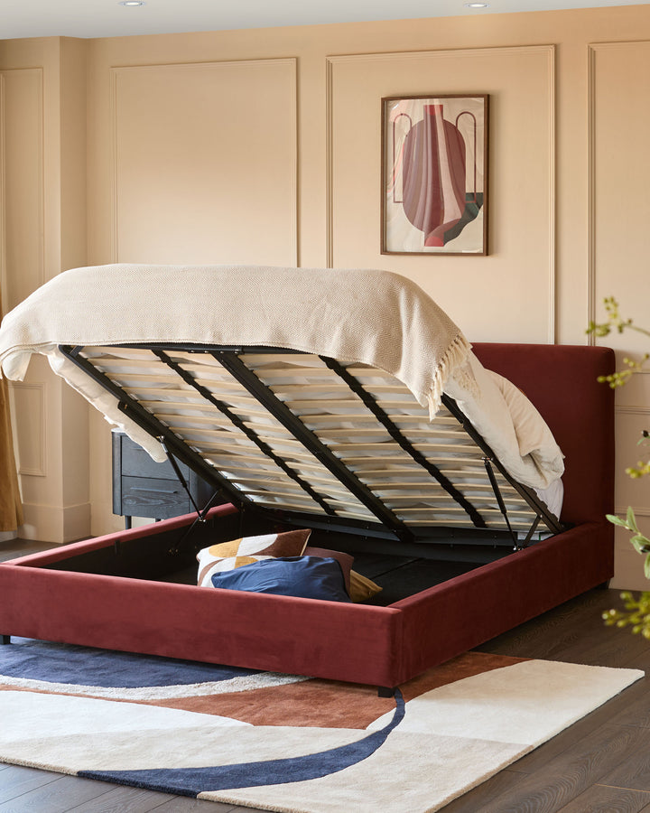 An upholstered bed in dark red with the mattress raised to reveal storage space underneath.