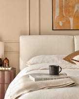 Bedroom scene with a cosy upholstered bed, wooden bedside table, book and mug. Warm and inviting interior.