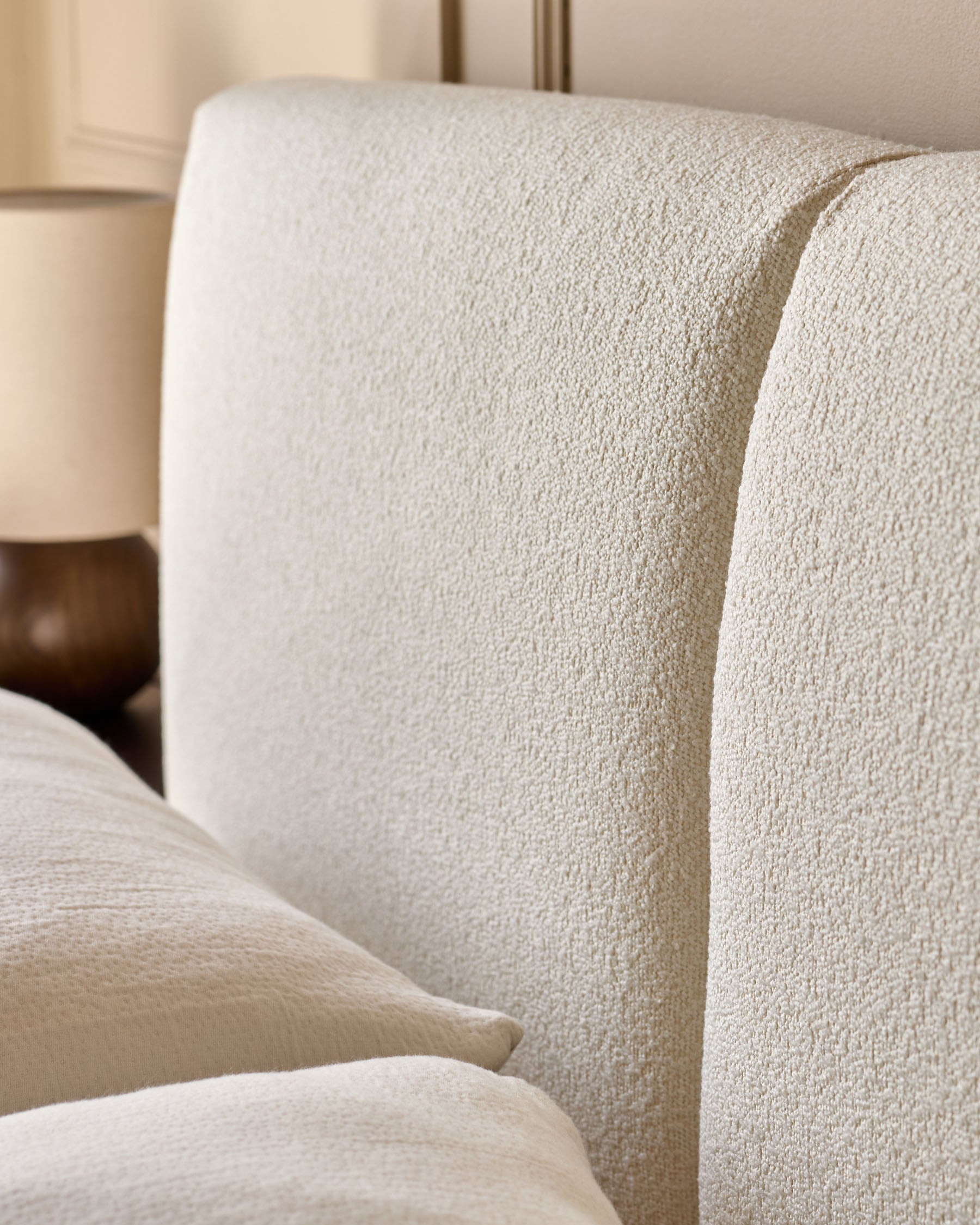 Close-up of a cream-coloured upholstered headboard and matching pillows with a bedside lamp in the background.