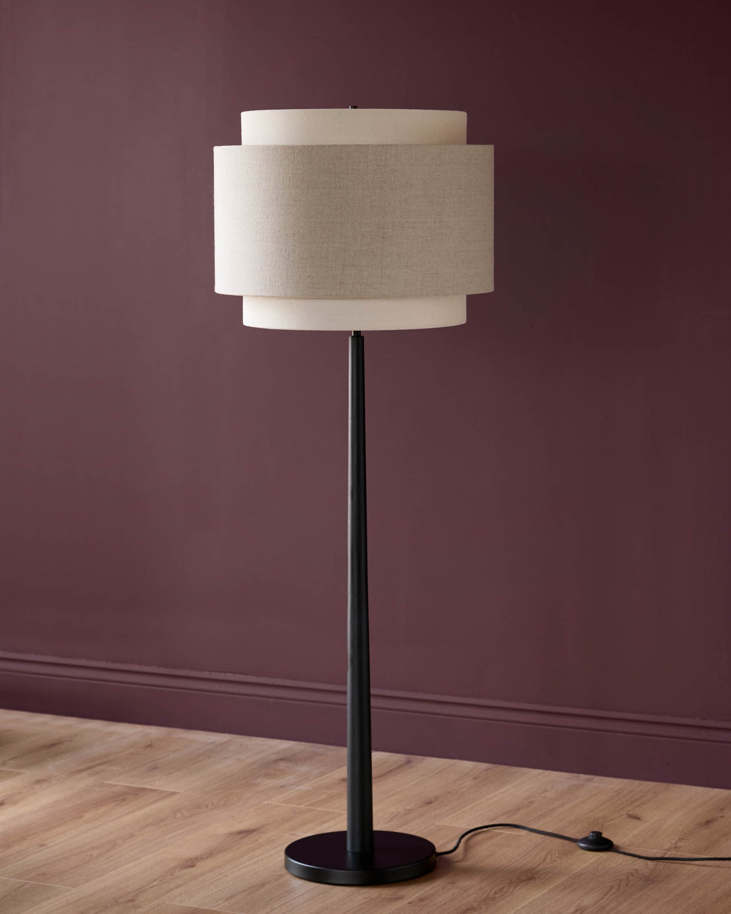 Leonie floor light with layered linen shade in natural and mocha tones, black metal stand and base, against burgundy wall.