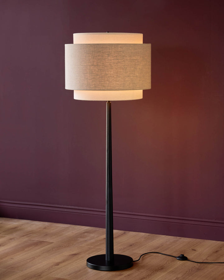 Leonie floor light with natural linen and mocha layered drum shade, dark metal pole base, illuminated against burgundy wall.