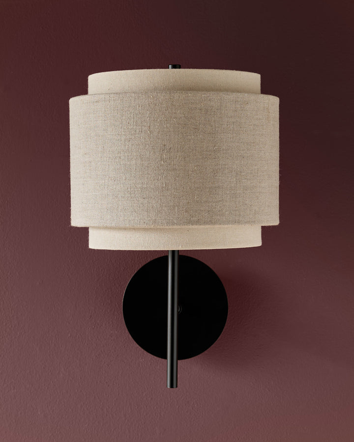 Leonie wall light with layered linen shades in natural and mocha tones, black circular backplate and stem against mauve background.