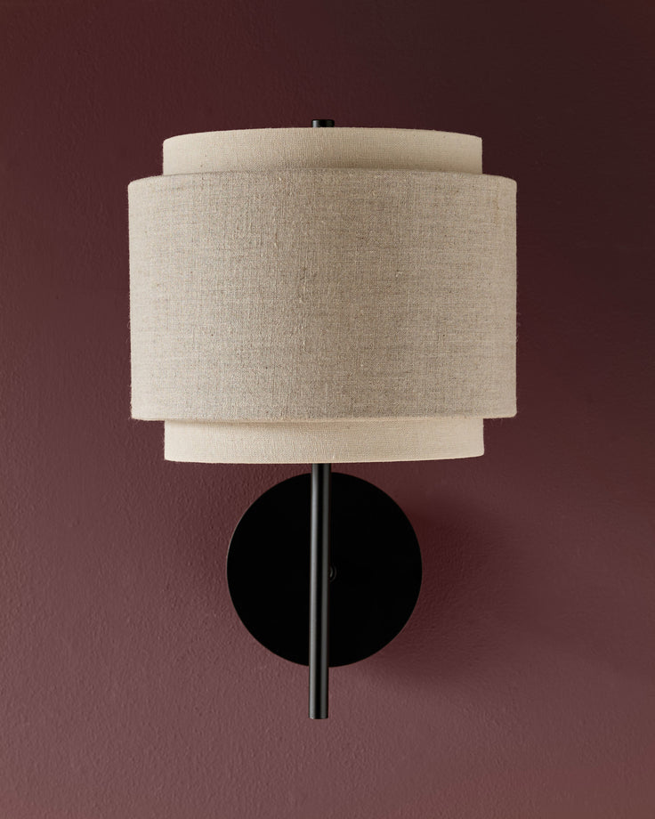 Leonie wall light with layered linen shades in natural and mocha tones, black circular backplate and stem against mauve background.