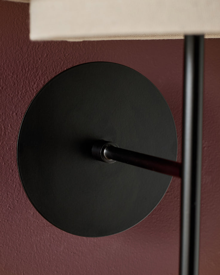 Black circular wall light with black arm mounted on burgundy textured wall, minimalist design detail.