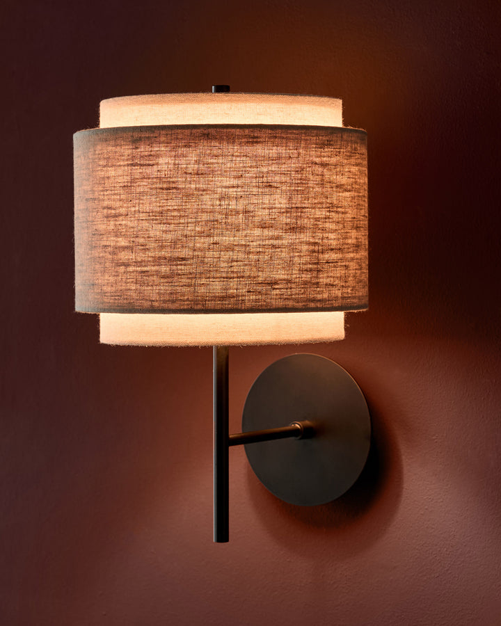 Leonie wall light with layered linen shade in natural and mocha, black metal fixture, mounted on brown wall.