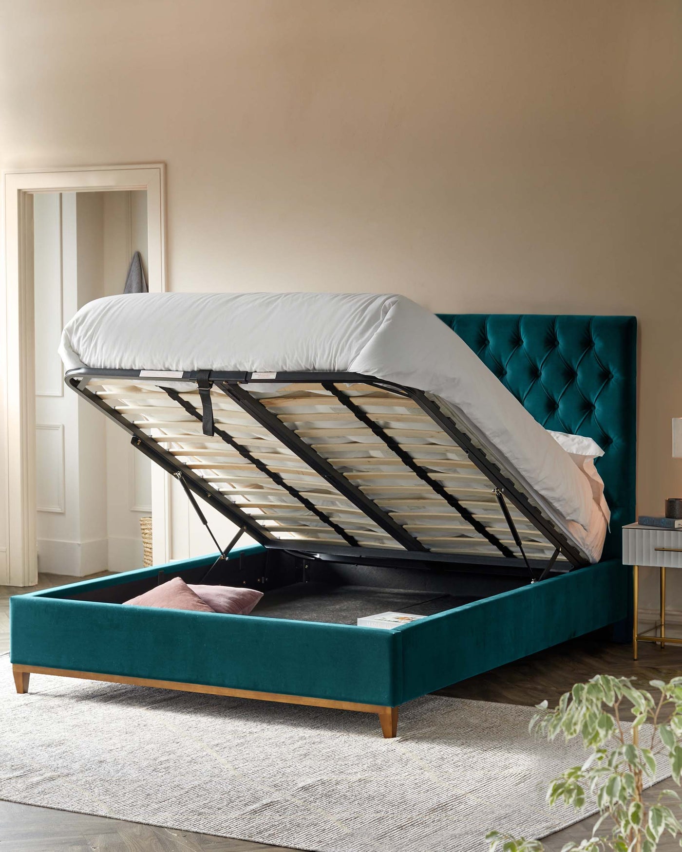 A luxurious teal upholstered storage bed with a lifted mattress platform revealing a spacious under-bed storage area. The bed features a tufted headboard and a wooden footing element.