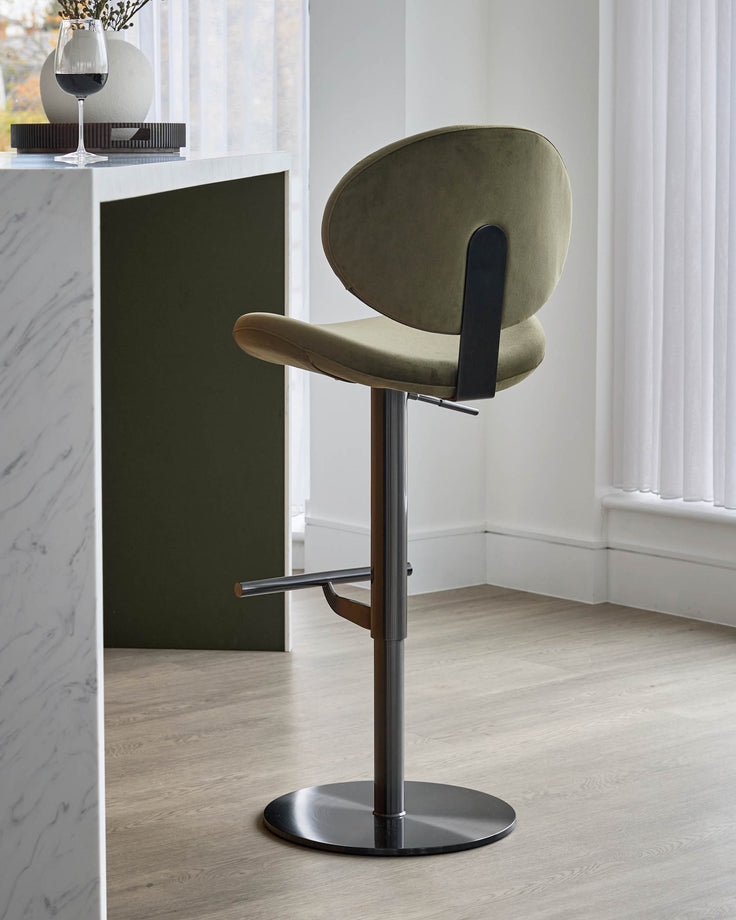 Lilah gas-lift bar stool in olive green velvet with black metal footrest and round base, shown in modern interior setting.