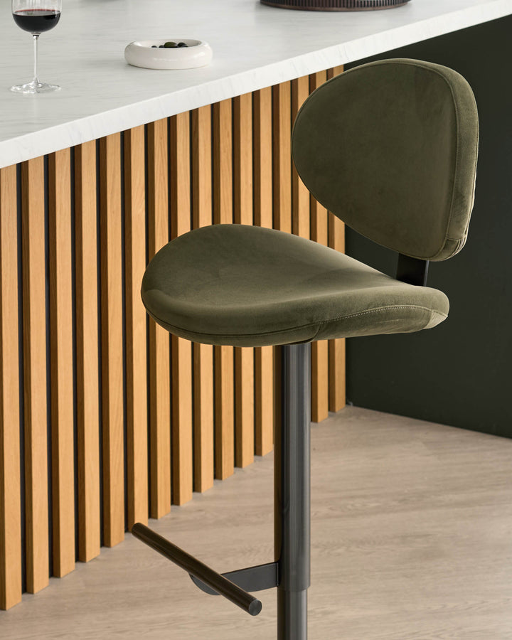 Lilah gas-lift bar stool in olive green velvet with rounded seat and backrest, shown against wooden slat paneling.