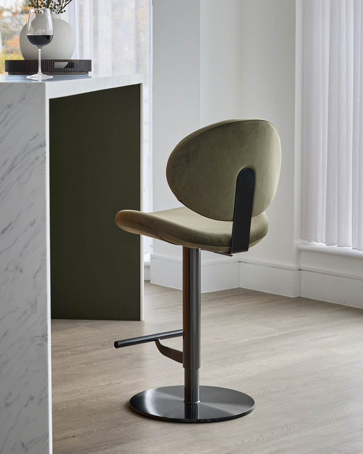 Lilah gas-lift bar stool in olive-green velvet with black metal base and footrest, shown in modern interior setting.