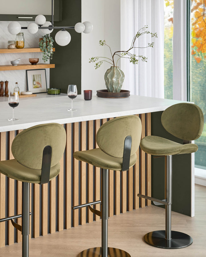 Lilah gas lift bar stool in olive green velvet with curved backrest and black metal base, shown in kitchen setting.