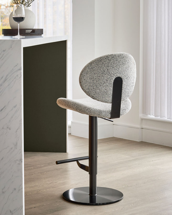 Lilah grey fabric gas lift bar stool with curved backrest, black metal base and footrest, shown in modern interior setting.