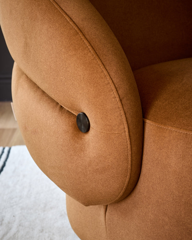 Caramel fabric accent chair detail showing curved armrest with black oval button accent and stitched seams.