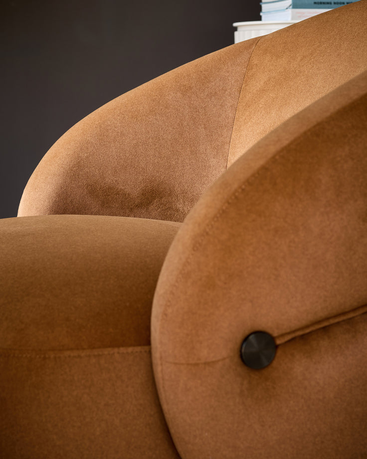 Lillian accent chair in caramel fabric, close-up of curved armrest and seat showing plush upholstery and black button detail.