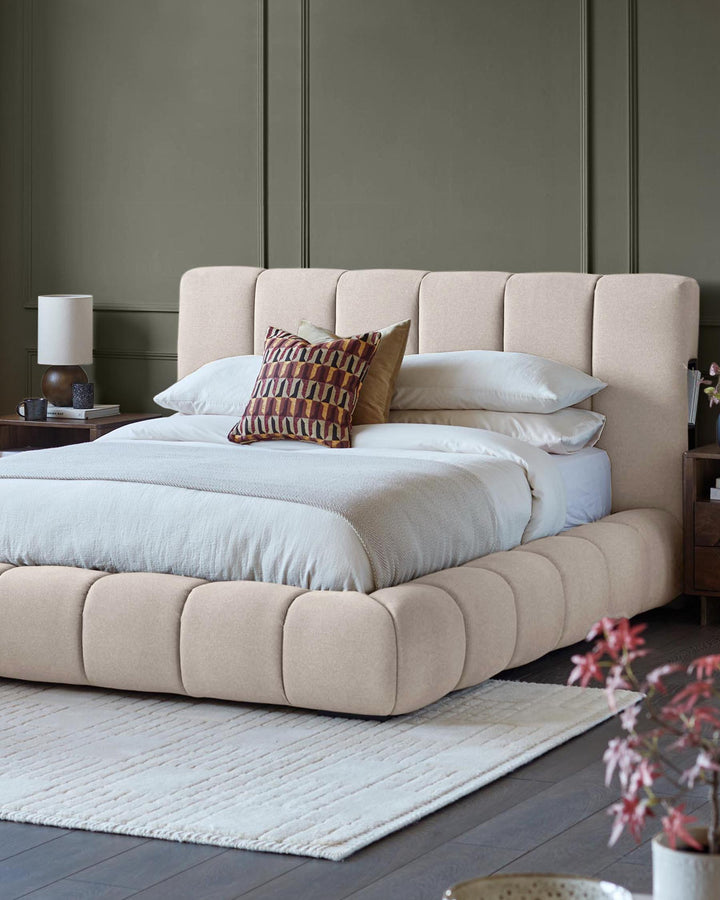 A padded beige platform bed with matching headboard, accessorised with patterned cushions and a cosy throw.