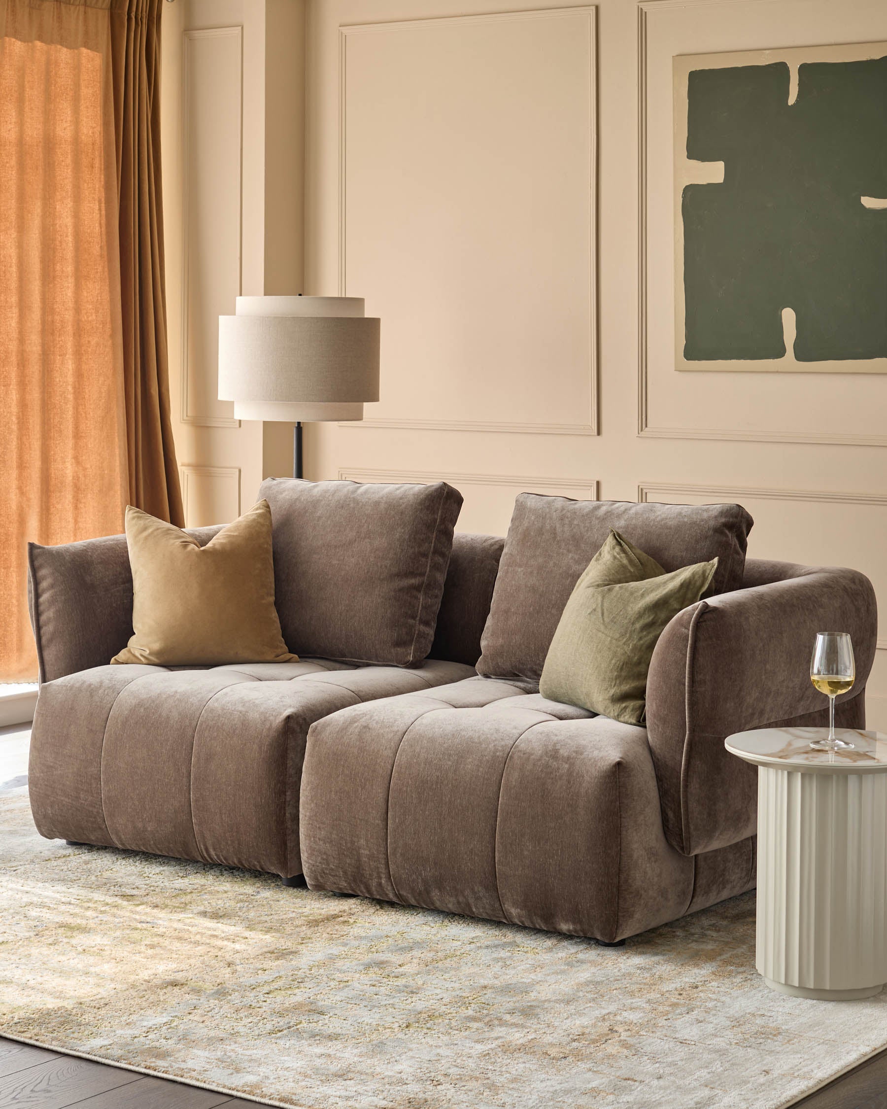 A contemporary living room features a brown sofa, neutral rug, side table, floor lamp, curtains and artwork.