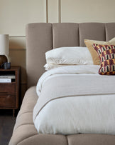 Lillian double ottoman bed in mocha fabric, shown from side angle with white bedding and patterned pillows, alongside wooden nightstand.