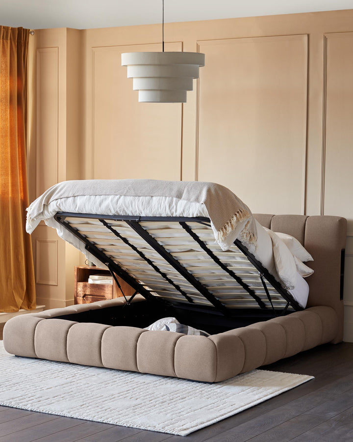 Upholstered ottoman bed in beige with lift-up storage, wooden side table, neutral walls and a cream rug.