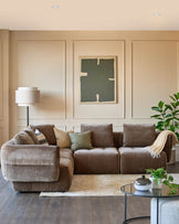 Stylish brown modular sofa with cushions, a round glass table, a floor lamp and artwork in a cosy living room setting.