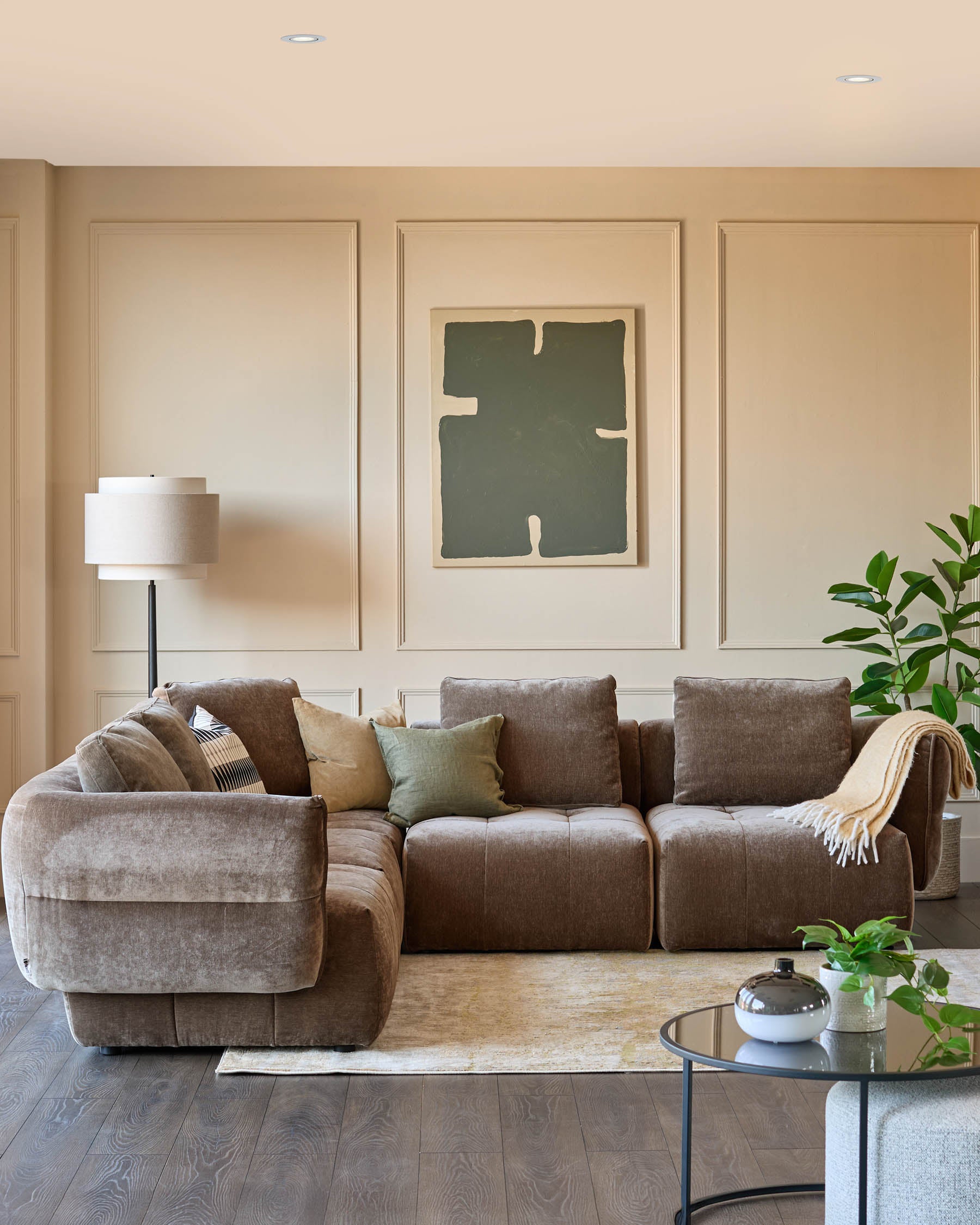 Stylish brown modular sofa with cushions, a round glass table, a floor lamp and artwork in a cosy living room setting.