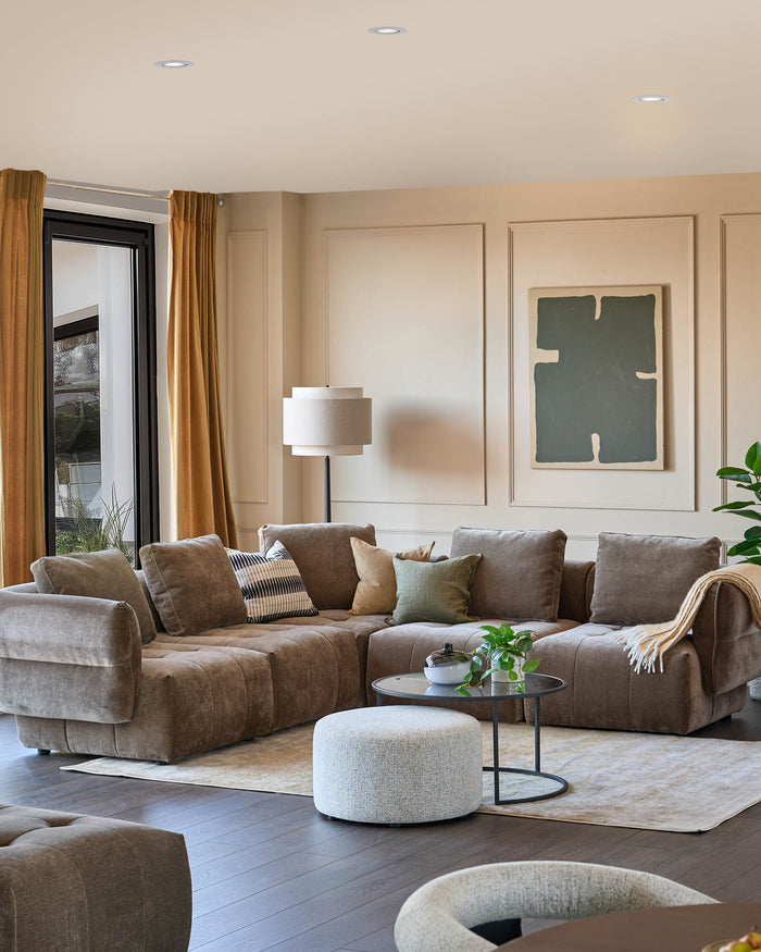 Living room featuring a modular sofa, round coffee table, footstool, floor lamp, & beige curtains, creating a cosy and stylish space.