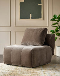 A plush brown armchair with a matching cushion, positioned on a patterned rug in front of a neutral wall and plant.