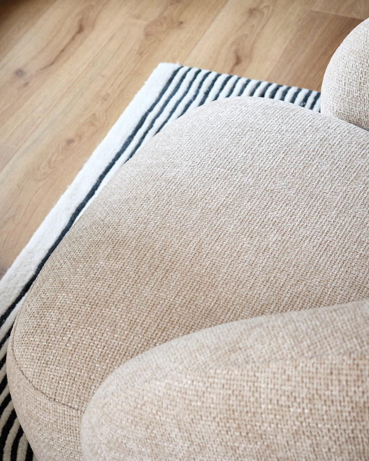 Lillian accent chair in natural fabric, shown from above at an angle highlighting the curved armrest and textured upholstery against hardwood flooring.