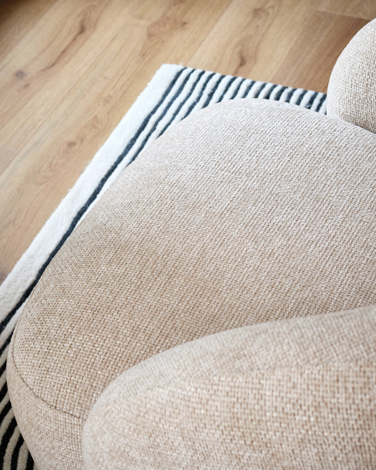 Lillian accent chair in natural fabric, shown from above at an angle highlighting the curved armrest and textured upholstery against hardwood flooring.