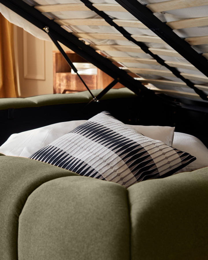 Olive-green fabric ottoman bed with striped pillow, angled view showing upholstered frame and open storage mechanism.