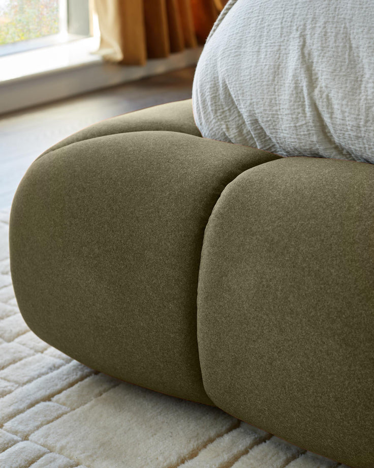 Olive green upholstered ottoman bed with rounded, curved design, shown in bedroom setting near window.