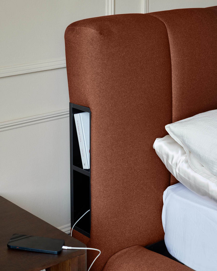 Toffee-red fabric ottoman bed with built-in storage compartment and charging port, shown with white bedding.