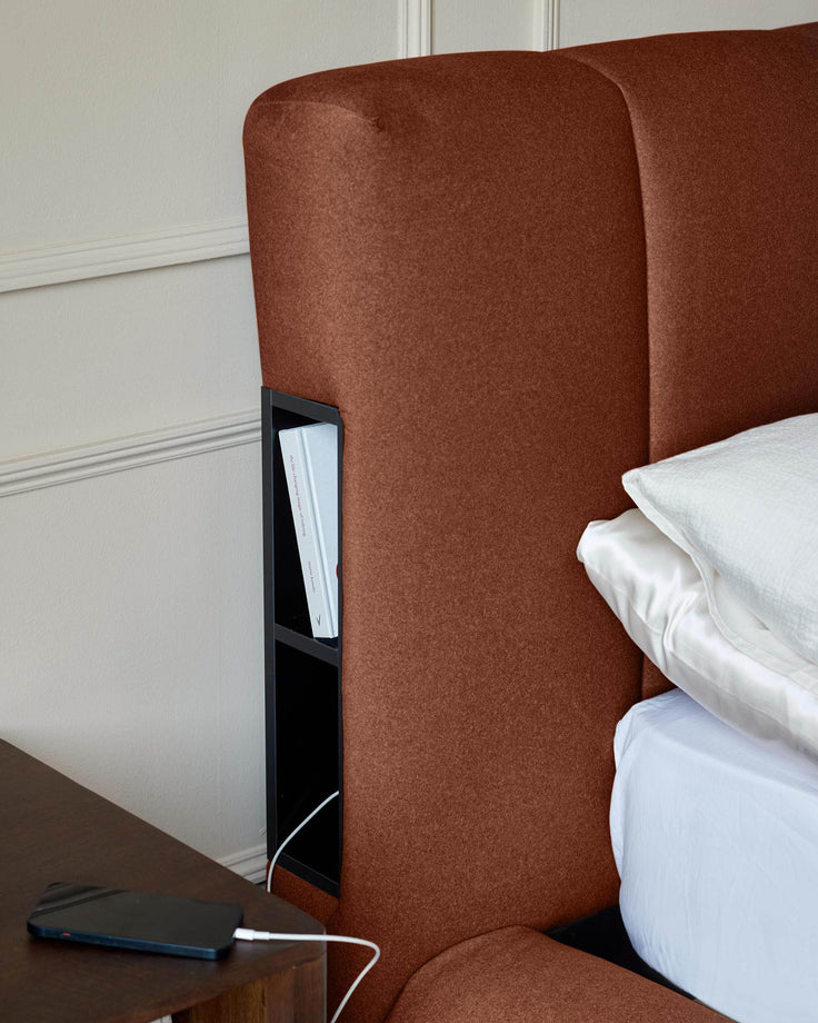 Toffee-red fabric ottoman bed with built-in storage compartment and charging port, shown with white bedding.