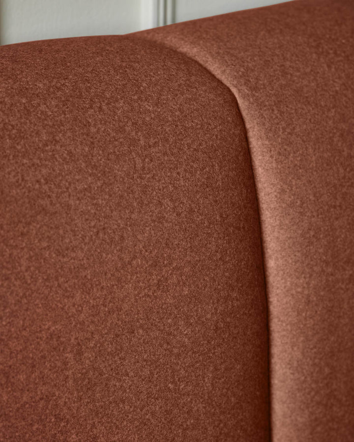 Toffee-red fabric ottoman bed showing curved headboard detail and upholstered surface texture.